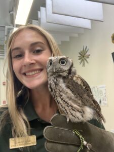 A person with a green shirt and gold name badge smiles at the camera while holding a small brown owl on a gloved hand.