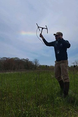 A person wearing brown chest waders, a blue shirt, and a baseball cap holds a radio receiver up to their ear as they walk across a field, holding up an H-shaped radio antenna with their other hand.