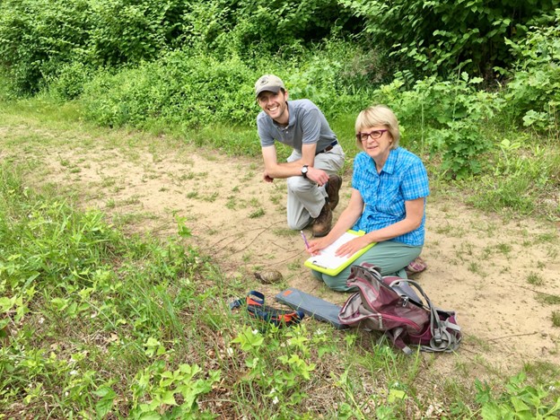 A man in a gray polo shirt, gray pants, and a ball cap kneels smiling beside a woman in a blue checked shirt and green pants. The woman has a clipboard on her lap and a radio with a long antenna in one hand. A box turtle sits on the ground in front of her in a large clearing/