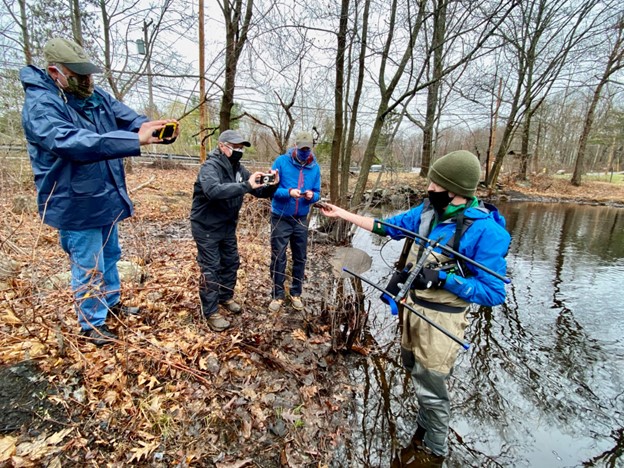 A man wearing a bright blue jacket, chest waders, a green knit cap, and a black face mask stands at the edge of a wide brook. He holds a radio antenna in one hand and is holding a small wood turtle in the other hand for three warmly-dressed people on shore to photograph with their phones. 