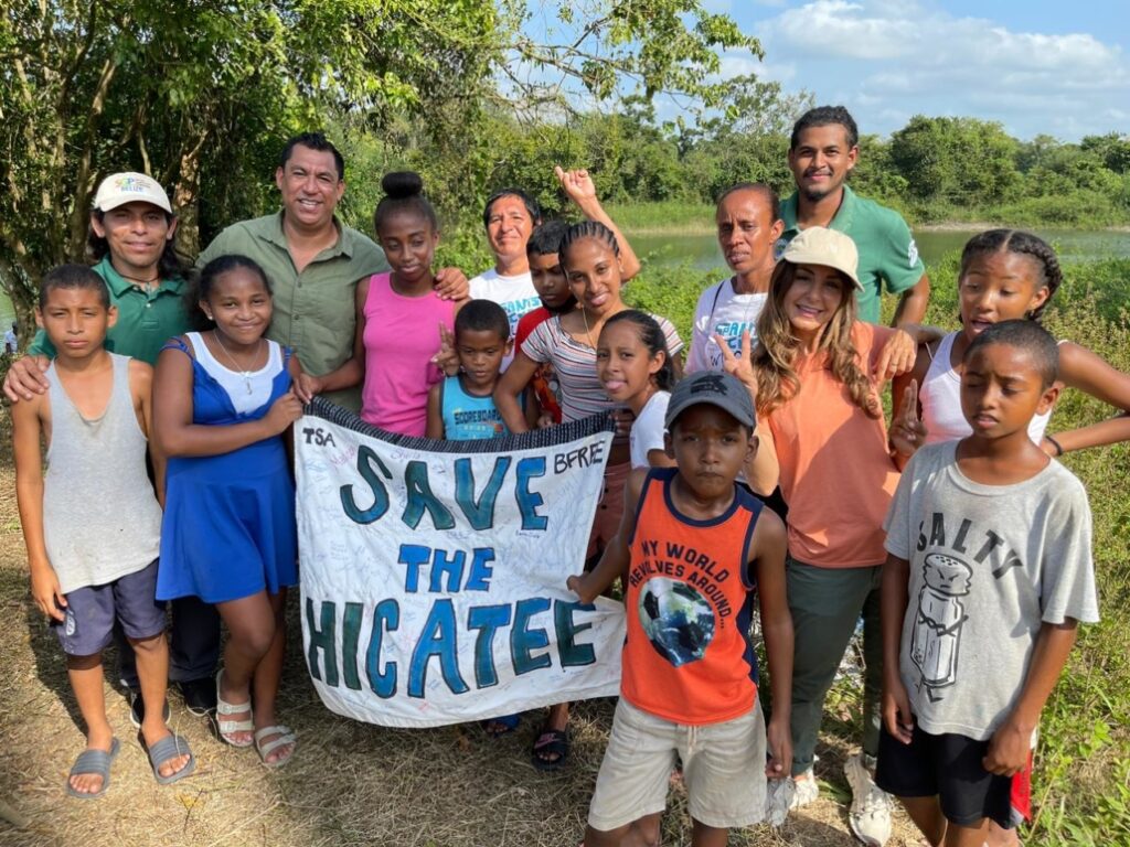 A crowd of smiling Belizean adults and children, including BFREE staff in green shirts, stand together on a sunny day with a wetland in the background. Two volunteers hold a homemade white banner with blue letters that reads, "Save the Hicatee"