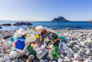 A pile of plastic garbage, mostly empty bottles, is piled up on a pebble beach with the ocean in the background.