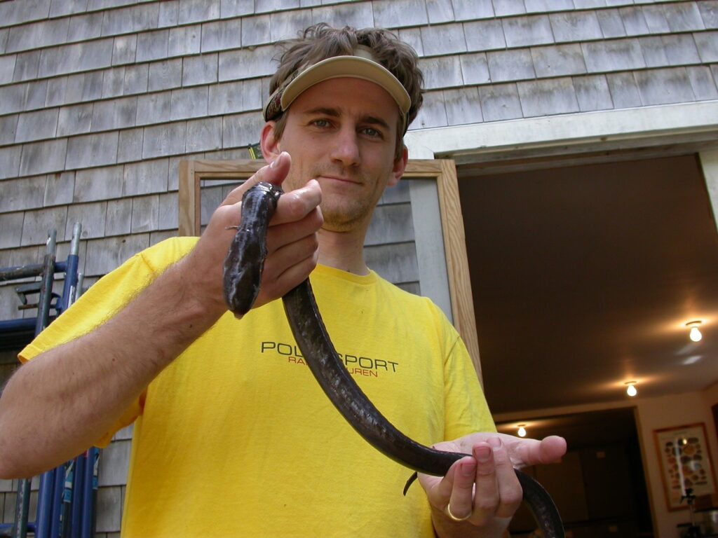 A man wearing a sun visor and a yellow t-shirt stands in front of a cedar-shingled building with an open door in the background. Draped between the man's hands is a large, dark, slimy amphibian with a long body and tiny vestigial forelegs.