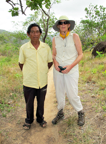 A man in a yellow shirt and black pants and a woman in a khaki vest, sun hat, and sunglasses stand side by side smiling at the camera against a backdrop of tropical scrubland.