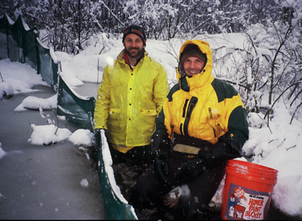 Two white men wearing yellow waterproof jackets and warm winter hats stand beside a green silt fence on the edge of a mostly-frozen pond. Snow covers the woods around them, and an orange five-gallon bucket sits beside them.