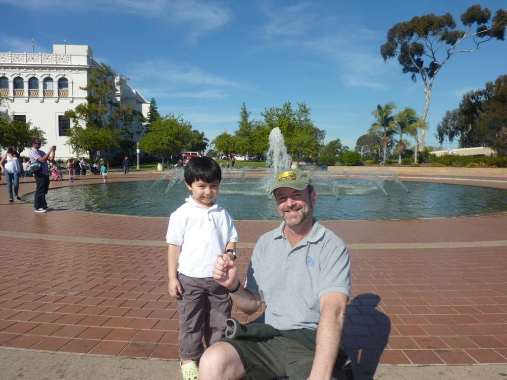 AA bearded man wearing a ball cap and a blue polo shirt kneels next to a dark-haired young boy in a white shirt. Both hold hands and smile at the camera in front of San Diego's Bea Evenson Fountain in Balboa Park.