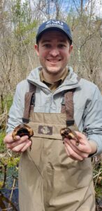 A person wearing chest waders and a ZNE blue ball cap smiles at the camera holding a spotted turtle in each hand.