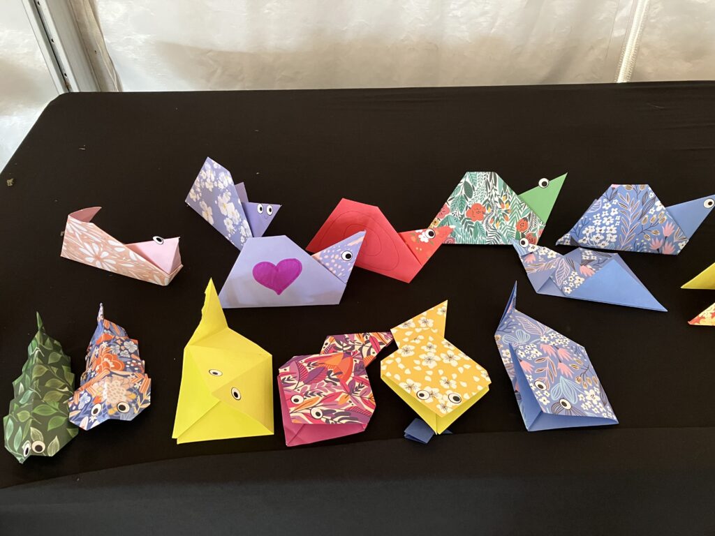 About a dozen colorful origami worms, snails, and tadpoles with attached googly eyes sit on a black tablecloth.