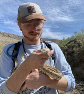 A person wearing a ball cap, a light-long-sleeved shirt, and glasses holds a snake carefully in his left hand against an arid backdrop.