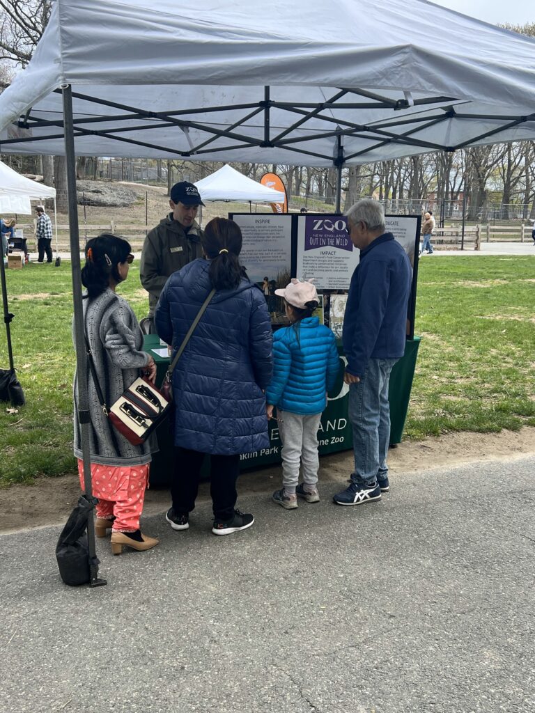 A man in a green coat with blue Zoo New England hat shows an interested family some information about ZNE's conservation efforts with handouts, pop-up displays, and a stuffed turtle.