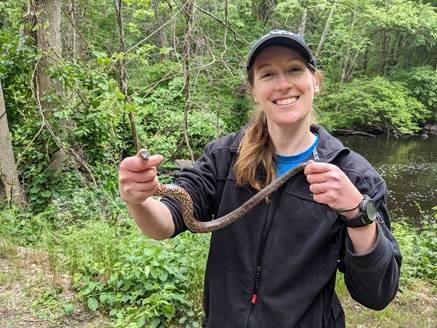 A smiling white woman with brown hair in a ponytail over her shoulder holds a brown snake between her hands while smiling at the camera. Behind her is a forested slope leading down to a river,