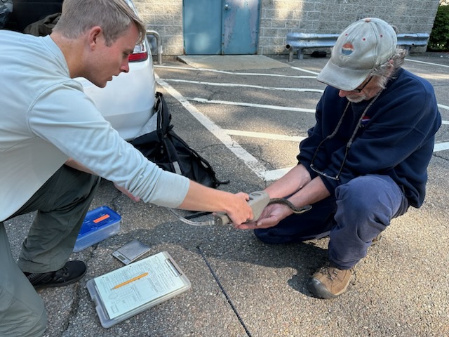 A young man with light skin and hair wearing a pale long-sleeved shirt holds a digital PIT tag reader over a snake held in the hands of an older light-skinned man with a white beard, a blue fleece top and cap with matching logo.