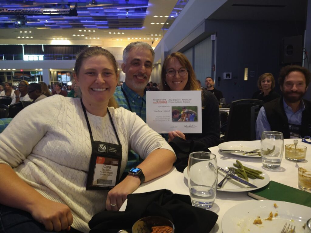 Four people seated around a table smile at the camera and hold up a square award saying: North American Conservation Award: Top Honors, Zoo New England.