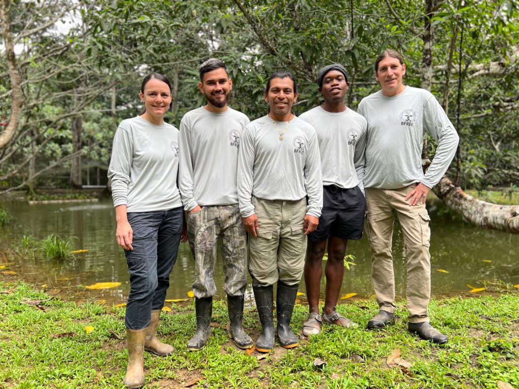 Five people in matching pale green shirts along with varied work pants and boots stand smiling side by side in front of a tropical wetland.