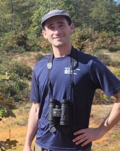 A white man with a blue ZNE hat and t -shirt wearing binoculars smiles at the camera against a backdrop of shrubs and trees with one hand on his hip.